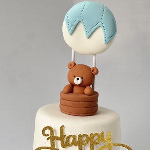 Fondant hot air balloon cake topper featuring a teddy bear in a basket, displayed on a white tiered cake with a gold “Happy Birthday” sign.