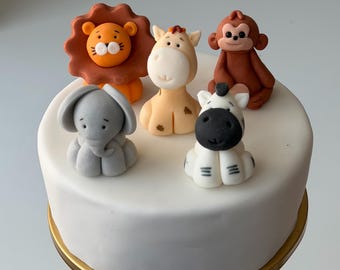 May include: A white frosted cake decorated with five animal figurines: a lion, giraffe, monkey, elephant, and zebra. The lion is orange, the giraffe is tan, the monkey is brown, the elephant is gray, and the zebra is white with black stripes.