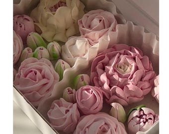 Op de afbeelding: Een doos gevuld met decoratieve, eetbare bloemen. De bloemen, in de vorm van rozen en tulpen, zijn in roze, witte en groene tinten. De opstelling bevindt zich in een witte doos met gekreukeld papier.