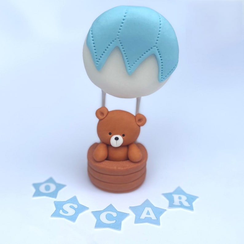 Edible cake topper made from sugar fondant, shaped as a brown teddy bear in a hot air balloon basket with a blue and white balloon. Below the topper, blue star-shaped fondant letters spell the name “Oscar”.