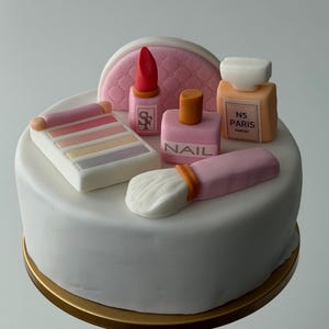 May include: A white cake decorated with fondant makeup items. The decorations include a pink lipstick, a nail polish bottle with "NAIL" text, a makeup brush, an eyeshadow palette, a perfume bottle with "N5 PARIS" text, and a pink quilted cosmetic bag.
