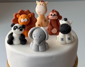 Può includere: Una torta glassata bianca decorata con figurine di animali in pasta di zucchero. Le figurine includono un leone, una giraffa, una scimmia, un panda, un elefante e una zebra. La torta ha una base dorata.