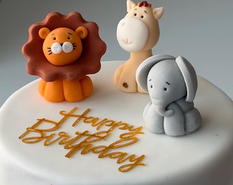 May include: A white frosted cake decorated with orange, brown, and gray animal figurines, including a lion, giraffe, and elephant. The cake is adorned with gold cursive text that reads "Happy Birthday".