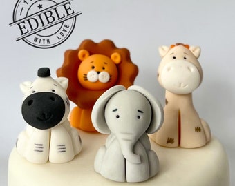 May include: Edible cake toppers featuring a zebra, lion, elephant, and giraffe. The lion has an orange mane and face. The zebra has black and white stripes. The elephant is gray, and the giraffe is tan. The toppers are on a white surface. The text "100% EDIBLE WITH LOVE" is in a circle.