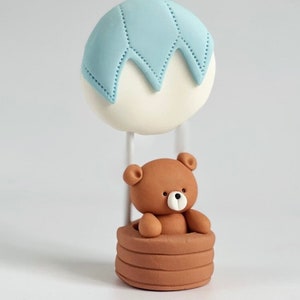 Fondant cake topper featuring a brown teddy bear sitting in a balloon basket with a light blue and white hot air balloon above.