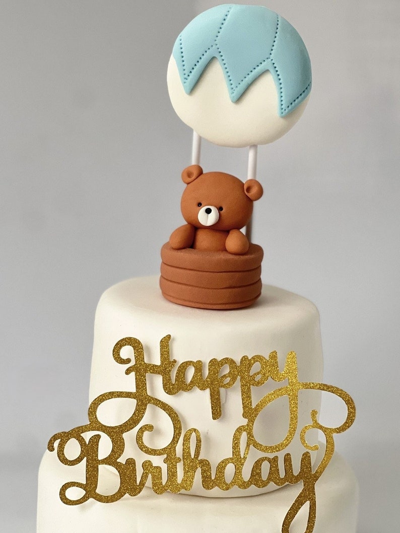 Fondant hot air balloon cake topper featuring a teddy bear in a basket, displayed on a white tiered cake with a gold “Happy Birthday” sign.