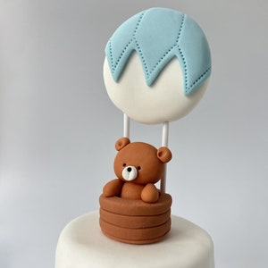 Edible cake topper made from sugar fondant, shaped as a brown teddy bear sitting in a hot air balloon basket with a white and light blue balloon. The topper is placed on a white frosted cake.