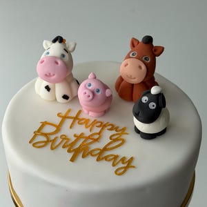 May include: A white iced cake featuring four farm animal cake toppers: a cow, a pig, a horse, and a sheep. The words "Happy Birthday" are written in gold script across the front of the cake.