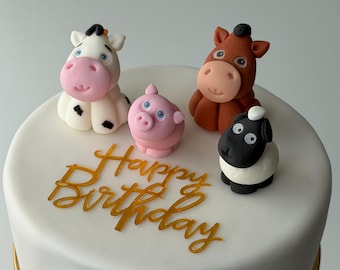 Pode incluir: Um bolo branco decorado com quatro figuras de animais de fazenda: uma vaca, um porco, um cavalo e uma ovelha. As palavras "Happy Birthday" estão escritas em letras douradas na frente do bolo.