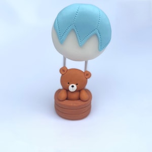 Edible cake topper made from sugar fondant, shaped as a brown teddy bear in a hot air balloon basket with a blue and white balloon.