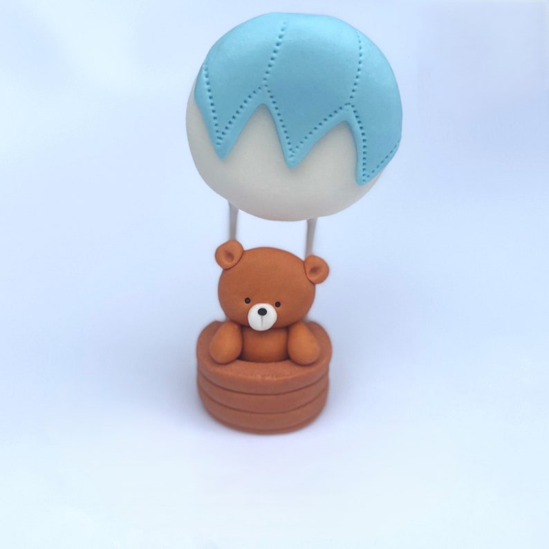 Edible cake topper made from sugar fondant, shaped as a brown teddy bear in a hot air balloon basket with a blue and white balloon.