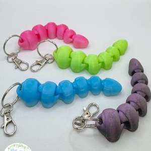 Puede incluir: Cuatro llaveros coloridos en rosa, verde, azul y morado. Cada llavero presenta un diseño segmentado en forma de gusano con un llavero y un cierre plateados. Los llaveros están dispuestos sobre una superficie blanca.
