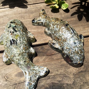 May include: Two resin fish sculptures with a silver and gold glitter finish. The fish are lying on a wooden surface.