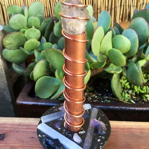 May include: A copper wire wrapped around a clear quartz crystal point, with a black base containing various crystals, including amethyst, selenite, and rose quartz.