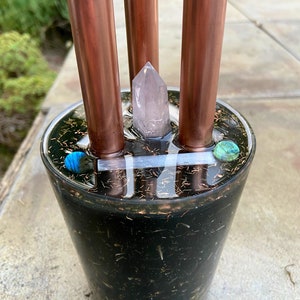 Op de afbeelding: Een zwarte hars wierookhouder met drie koperen pijpen en een helder kristal in het midden. De houder is gevuld met zwart zand en heeft een kleine turquoise steen aan de rand.