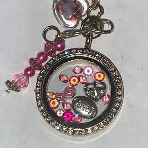 May include: A gold locket necklace with a pink ribbon charm, a small heart charm, and the word "faith" inside. The locket is filled with pink and orange crystals.