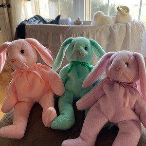 May include: Three plush bunny toys, one pink, one light green, and one lavender, sitting on a brown surface.