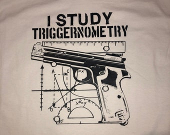May include: Cream-colored t-shirt with the text "I STUDY TRIGGERNOMETRY" in bold black letters. A detailed black graphic of a handgun is overlaid with geometric diagrams and a ruler, creating a unique design.