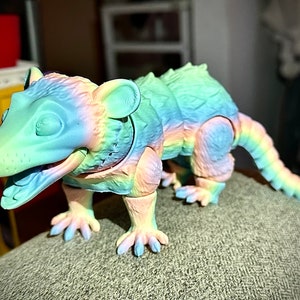 May include: A rainbow-colored 3D printed opossum figurine with a textured surface. The opossum is posed in a walking position with its mouth open and its tail curled behind it.