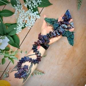 May include: A colorful, iridescent, dragon-shaped bracelet made of polymer clay. The bracelet is wrapped around a person's arm and features a variety of shades of blue, green, and purple.