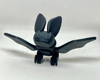 Könnte beinhalten: Eine schwarze Plastik-Fledermaus-Figur mit großen Ohren und Flügeln. Die Fledermaus hat ein einfaches, cartoonartiges Gesicht.