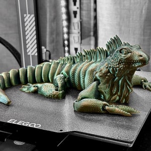 Op de afbeelding: Een 3D-geprint groen leguaan-hagedismodel op een zwart en grijs textuurvlak met het woord "ELEGOO" in wit gedrukt.