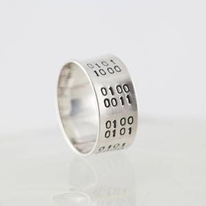 Puede incluir: Un anillo de plata con c&oacute;digo binario grabado en la banda. El c&oacute;digo dice "0101 1000 0100 0011 0100 0101".