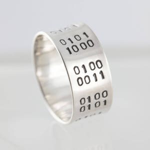 Puede incluir: Un anillo de plata con c&oacute;digo binario grabado en la banda. El c&oacute;digo dice "0101 1000 0100 0011 0100 0101".