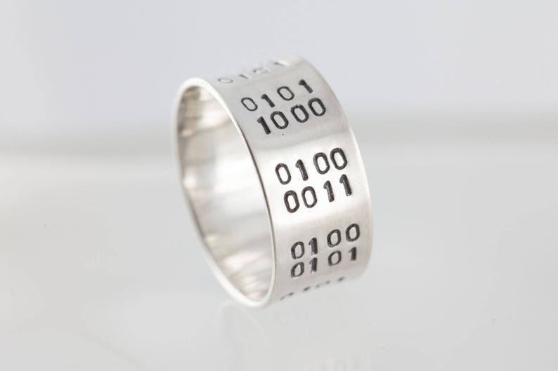 Puede incluir: Un anillo de plata con c&oacute;digo binario grabado en la banda. El c&oacute;digo dice "0101 1000 0100 0011 0100 0101".