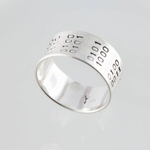 Puede incluir: Un anillo de plata con un patr&oacute;n de c&oacute;digo binario grabado alrededor de la banda.
