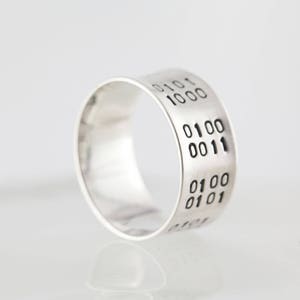 Puede incluir: Un anillo de plata con una banda ancha grabada con c&oacute;digo binario: 0101 1000 0100 0011 0100 0101.