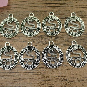 May include: Eight silver-toned metal charms featuring a snake design. Each charm depicts a snake coiled within a circular frame with a geometric pattern.