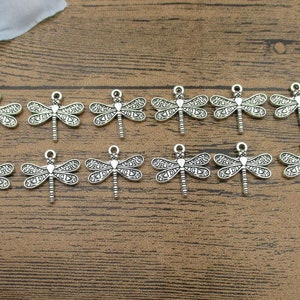 Op de afbeelding: Een set van 14 zilverkleurige libellenbedels. De bedels zijn gedetailleerd met ingewikkelde patronen en hebben een lus aan de bovenkant om aan sieraden of andere knutselwerkjes te bevestigen.