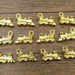 May include: A collection of small, gold-colored train charms. Each charm features a detailed locomotive design with wheels, a smokestack, and a loop for attaching to jewelry or other crafts. The charms are arranged on a wooden surface.