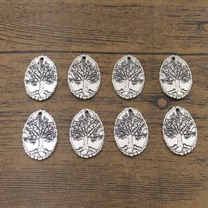 Peut inclure: Huit pendentifs ovales argentés, chacun avec un motif d'arbre détaillé. Les pendentifs ont un petit trou en haut pour l'enfilage. Le motif de l'arbre comprend des racines, un tronc, des branches et des feuilles.