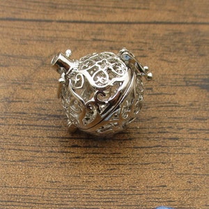 May include: Silver filigree cage charm with a delicate design. The cage is open on one side and has a small loop at the top for attaching to a necklace or bracelet.