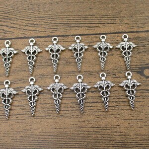 May include: A collection of silver-colored Caduceus charms, a symbol of medicine, arranged on a wooden surface. Each charm features a winged staff with two snakes entwined around it, and a small loop at the top.