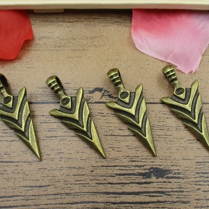 May include: Four antique gold-toned arrowhead pendants with a detailed design. The arrowheads are about 2 inches long and have a loop at the top for attaching to a chain or cord.
