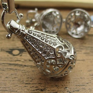 Könnte beinhalten: Eine silberne Charm-Kette mit einem detaillierten, verzierten Anhänger. Der Anhänger ist tropfenförmig und hat ein zartes, filigranes Design. Die Kette ist perfekt, um jedem Outfit einen Hauch von Eleganz zu verleihen.