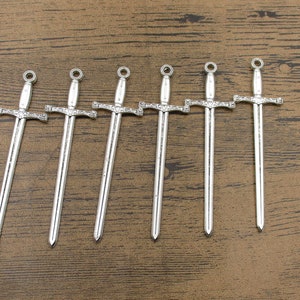 May include: Six silver sword charms with ornate details. The swords are approximately 5 cm long and have a loop at the top for attaching to a necklace or bracelet.
