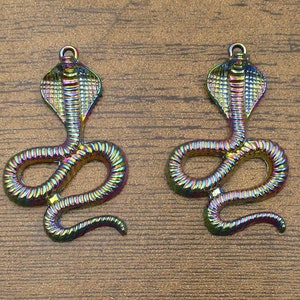 Op de afbeelding: Twee regenboogkleurige metalen slangencharms met een cobrakopontwerp. De charms zijn in de vorm van een opgerolde slang met een opgeheven kop.