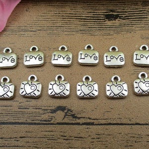 Puede incluir: Diez colgantes de plata con un corazón o la palabra "love" grabada en ellos. Los colgantes son todos del mismo tamaño y forma, y están dispuestos en dos filas de cinco.