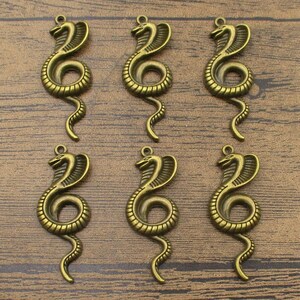 Op de afbeelding: Zes antieke goudkleurige slangbedels met een opgerold lichaam en een opgeheven kop. De bedels zijn perfect voor het maken van sieraden of knutselen.