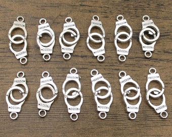 Pode incluir: Doze pingentes de metal prateados em forma de algemas com a palavra "FREEDOM" gravada em cada um. Os pingentes têm dois laços de cada lado para serem presos a joias ou outros artesanatos.