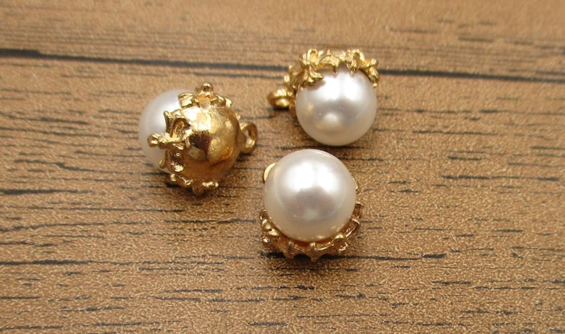 Puede incluir: Tres botones de perla blanca con adornos de metal dorado. Los botones tienen un dise&ntilde;o decorativo con un motivo floral.