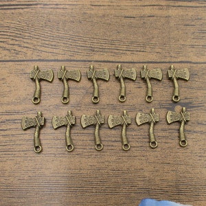 Pode incluir: Um conjunto de 12 pingentes de machado de cor dourada antiga. Os pingentes são detalhados com desenhos intrincados e têm um laço na parte superior para serem presos a joias ou outros artesanatos.
