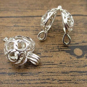 Może przedstawiać: Dwa srebrne wiszące elementy w kształcie klatki z filigranowymi zdobieniami, z pętelką w kształcie łzy u góry. Wiszące elementy są otwarte i mają delikatny, misterny wzór.