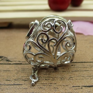 Könnte beinhalten: Silberner filigraner herzförmiger Käfiganhänger mit einer kleinen Kette, die an der Unterseite befestigt ist.