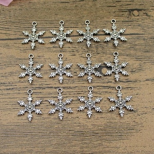 Op de afbeelding: Twaalf zilveren sneeuwvlokcharms met een gedetailleerd ontwerp. Elke charme heeft een lus aan de bovenkant om te bevestigen aan sieraden of andere knutselprojecten.