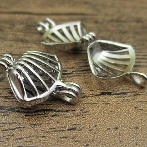 Peut inclure: Deux cages à perles en métal argenté en forme de coquillages. Chaque pièce présente une conception articulée avec une structure ajourée nervurée et une petite boucle en haut et en bas pour la fixation, permettant d'ouvrir et de fermer la cage.